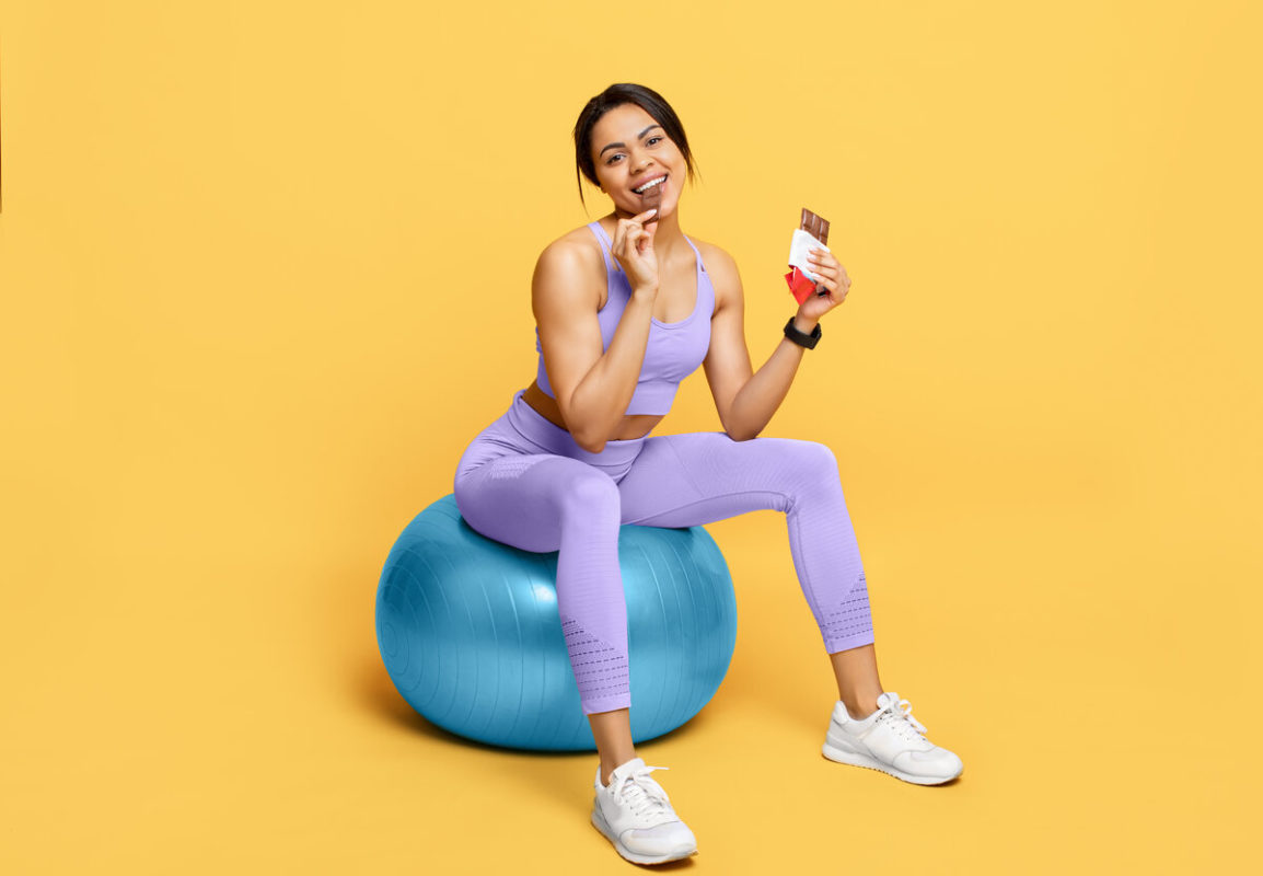 Happy african american woman sitting on fitball and eating chocolate, having break after training, yellow background eating sugar-free chocolate