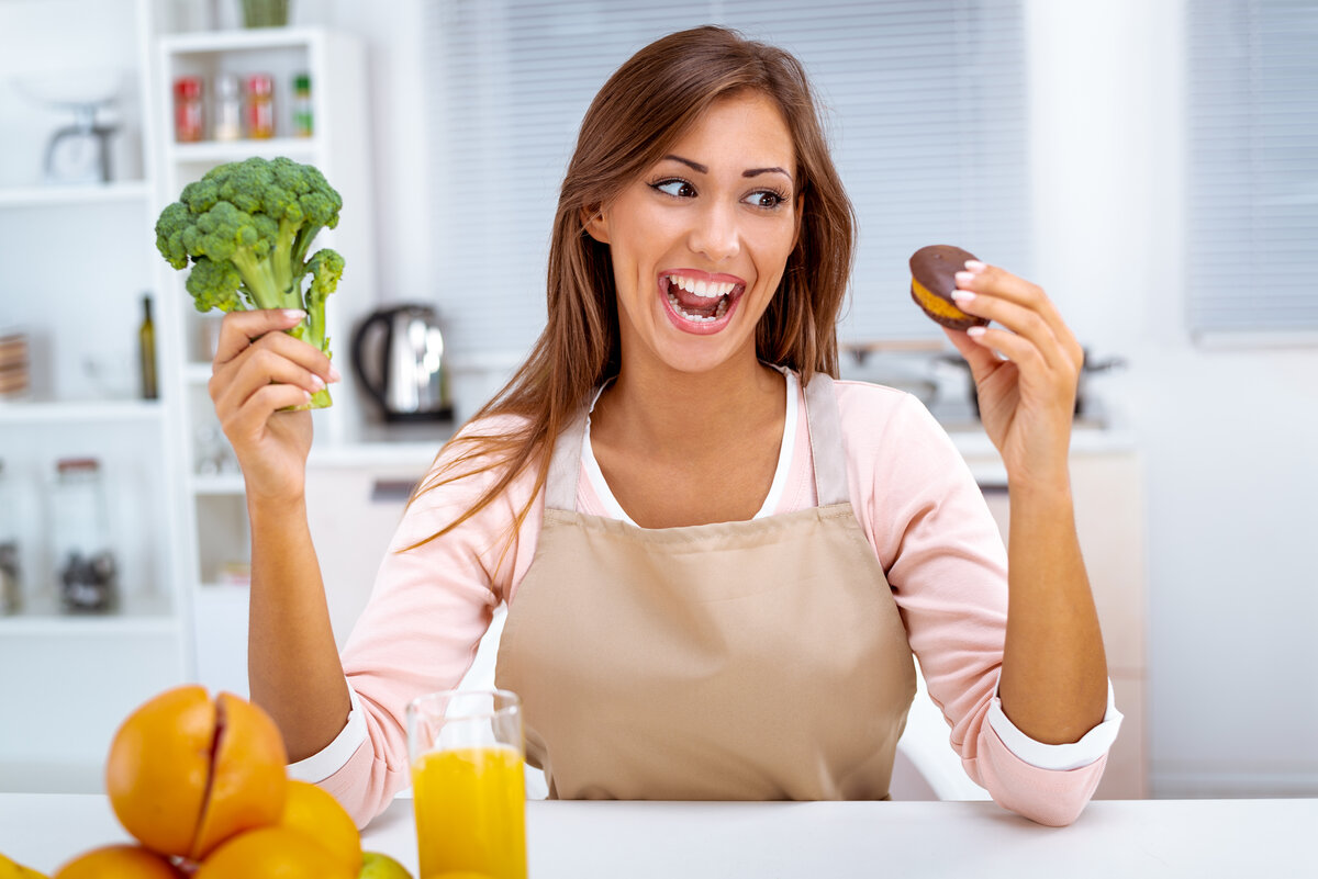 Beautiful young woman in the kitchen decision between broccoli and sweet chocolate cake for dieting