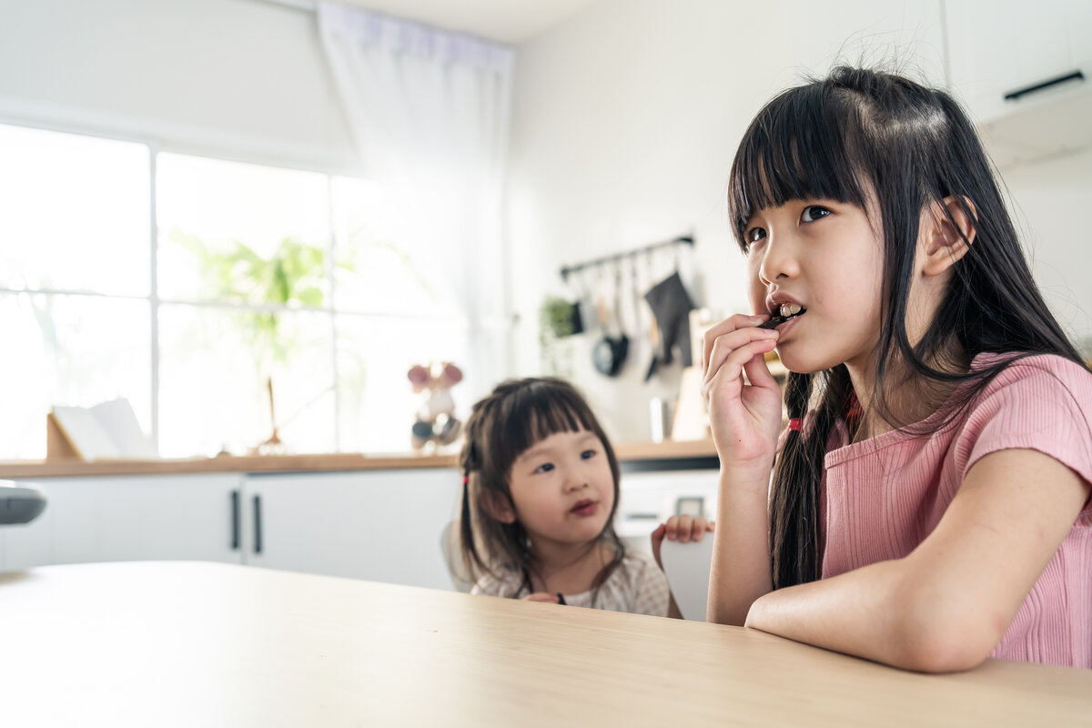 Portrait of Asian happy little cute kid holding chocolate in kitchen. Young lovely preschool child sibling sister sit on table enjoy eating sweet chocolate bar and smiling, looking at camera at home.