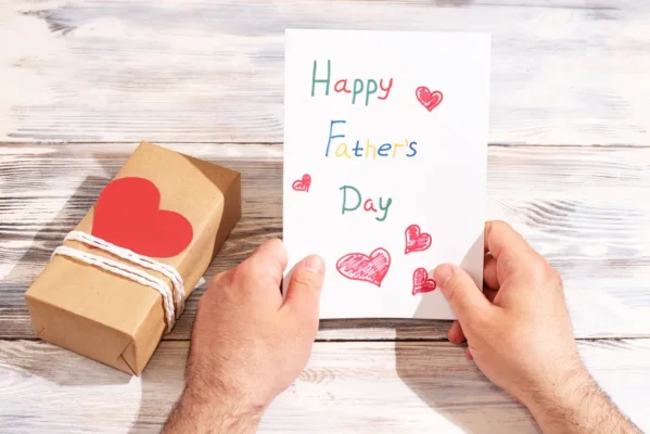 Male hands holding a greeting card made by a child on a white wooden background with a gift, congratulations to father - Father's Day Gift
