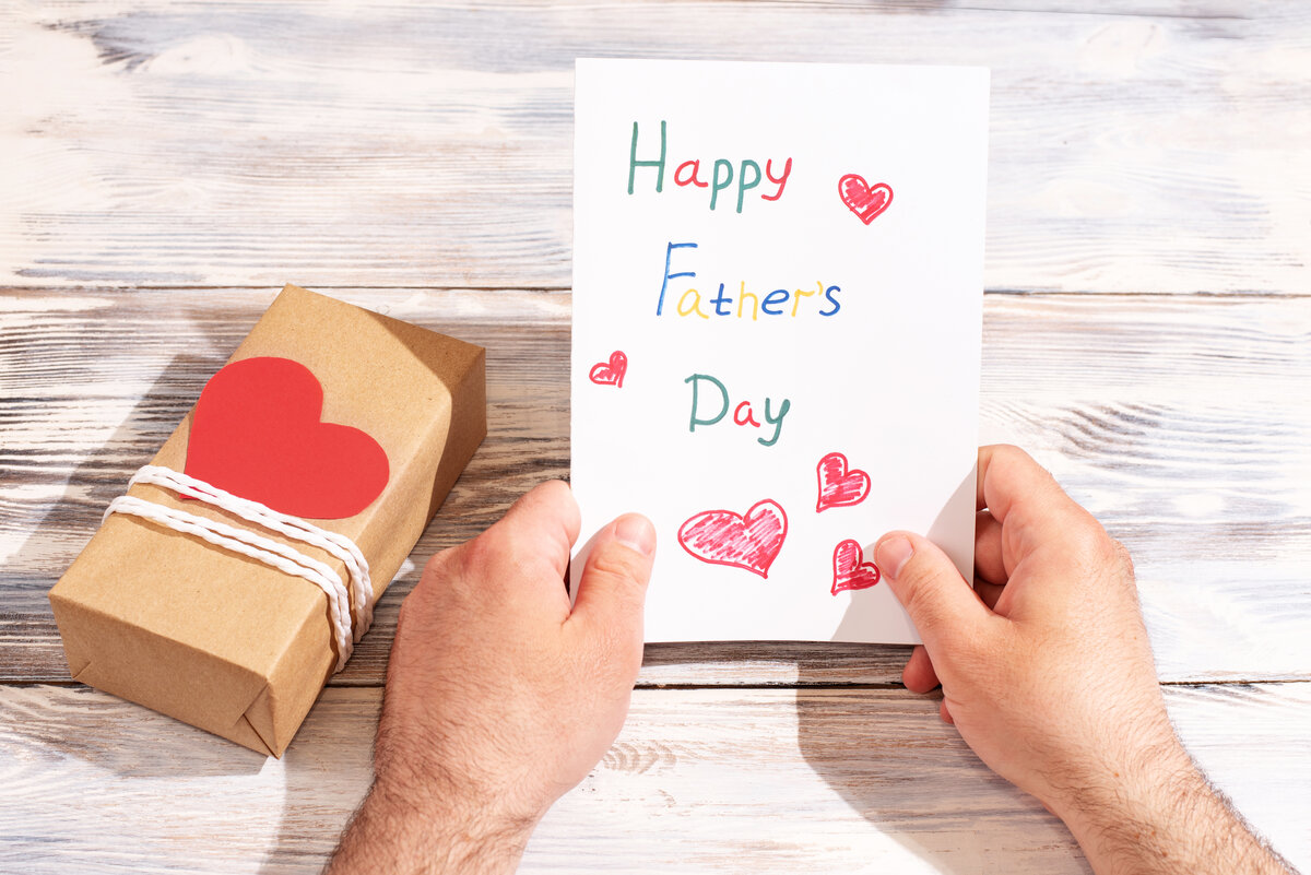 Male hands holding a greeting card made by a child on a white wooden background with a gift, congratulations to father - Father's Day Gift